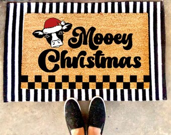May include: A rectangular doormat with a black and white striped border. The center features a cow wearing a Santa hat and the words "Moovey Christmas" in black script. A black and white checkered pattern is at the bottom.