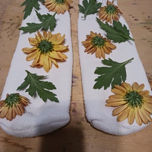May include: Two white socks with yellow flowers and green leaves arranged on them. The socks are laid out on a wooden surface.