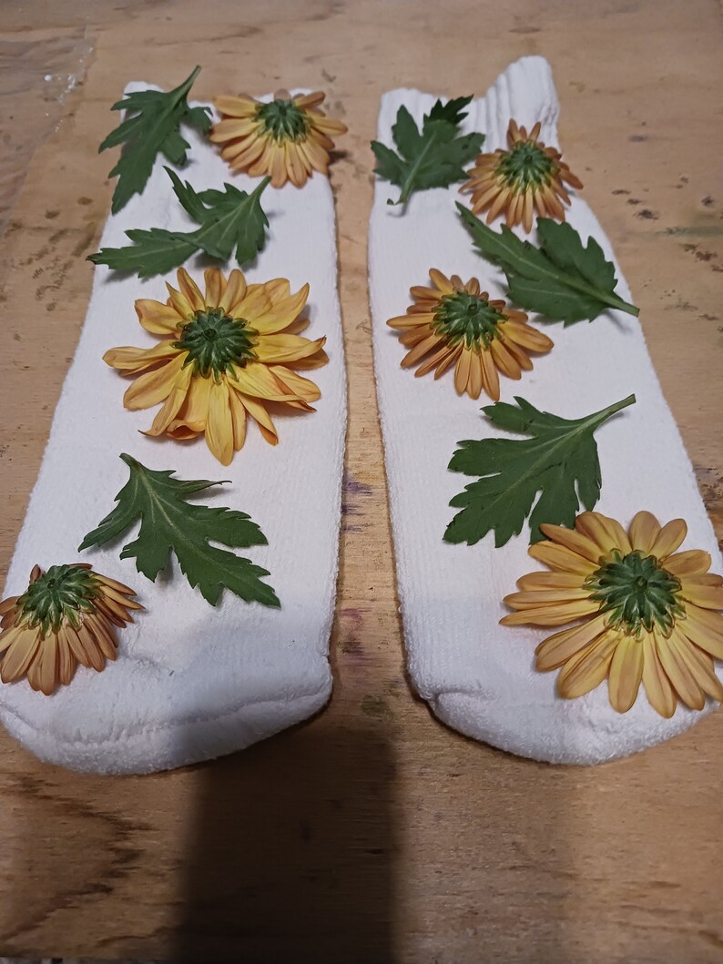 May include: Two white socks with yellow flowers and green leaves arranged on them. The socks are laid out on a wooden surface.
