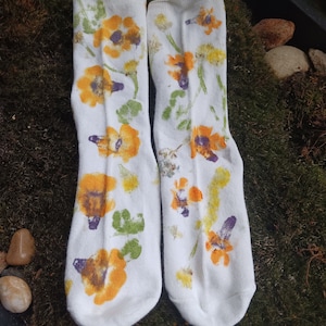 May include: A pair of white socks with a floral print. The flowers are yellow, orange, and purple. The socks are on a green mossy surface.