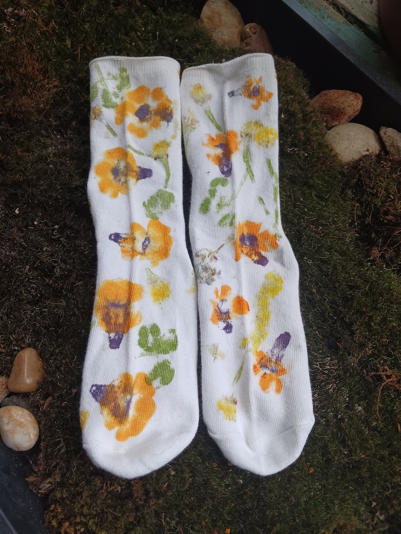 May include: A pair of white socks with a floral print. The flowers are yellow, orange, and purple. The socks are on a green mossy surface.
