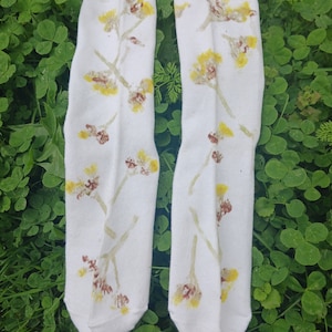 May include: A pair of white socks with a floral pattern in shades of yellow and brown. The socks are laid out on a bed of green clover.