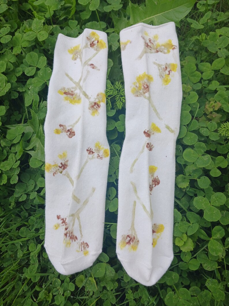 May include: A pair of white socks with a floral pattern in shades of yellow and brown. The socks are laid out on a bed of green clover.