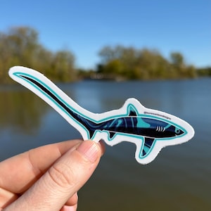 May include: A sticker featuring a stylized thresher shark design. The shark is primarily dark blue with teal and black accents, outlined in teal. The sticker has the text "@laurencarlsondesigns".