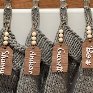 May include: Four gray knit Christmas stockings hang from a wooden rod. Each stocking has a wooden tag with a name in white script and a string of wooden beads. One tag has a paw print.