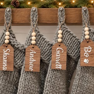 May include: Four gray chunky knit Christmas stockings hanging in a row. Each stocking features a wooden tag attached with twine and three light-colored wooden beads. The tags display the white names Shanna, Andrew, Garrett, and Bo with a paw print.