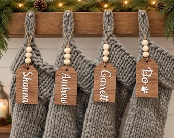 May include: Four gray chunky knit Christmas stockings hanging in a row. Each stocking features a wooden tag attached with twine and three light-colored wooden beads. The tags display the white names Shanna, Andrew, Garrett, and Bo with a paw print.
