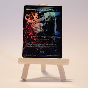 Op de afbeelding: Een ruilkaart getiteld "Bloodthirsty Conqueror" met een illustratie van een vrouwelijke vampierridder. De kaart heeft een zwarte rand en tekst die de vaardigheden en statistieken van het wezen beschrijft. De kaart wordt gepresenteerd op een klein houten ezel.