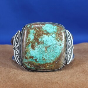 May include: A turquoise and silver cuff bracelet. The large, square turquoise stone has brown matrix patterns. The silver cuff features decorative elements on each side. The background is a solid blue.