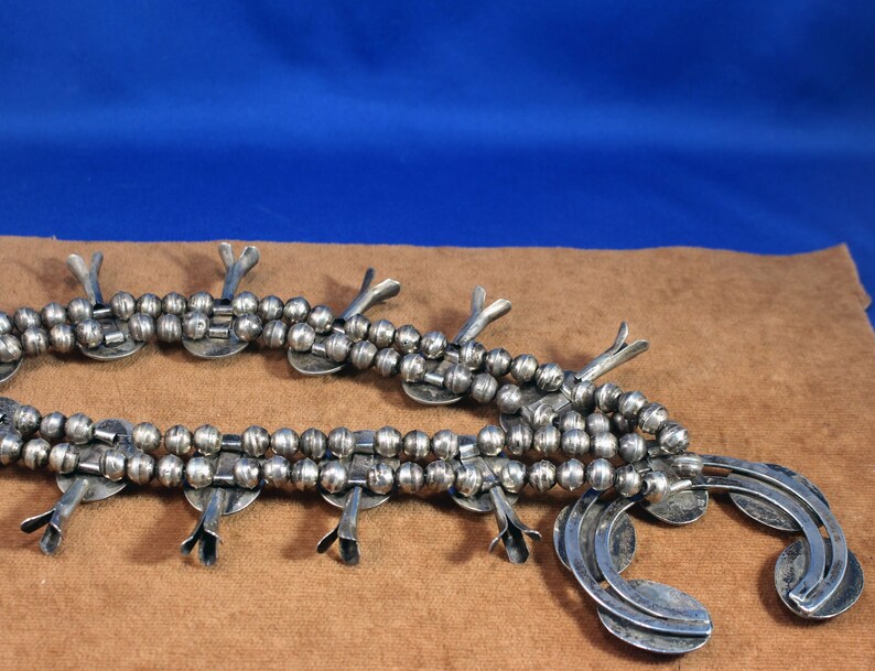 May include: A silver Native American squash blossom necklace with a beaded chain and multiple silver pendants. The necklace is laid out on a brown surface.