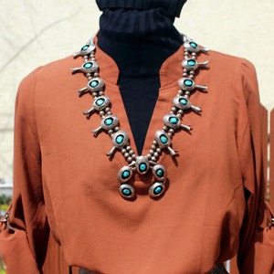 May include: A rust-coloured blouse with a black turtleneck underneath. The blouse has a silver and turquoise necklace with a southwestern design.