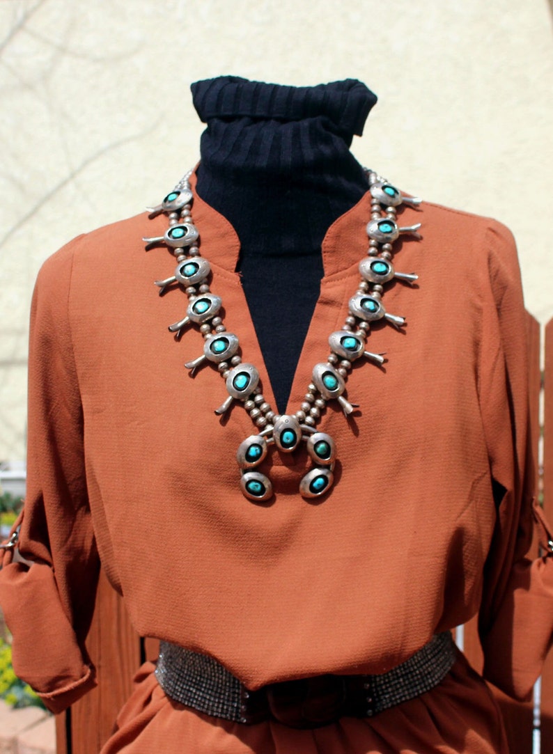 May include: A rust-coloured blouse with a black turtleneck underneath. The blouse has a silver and turquoise necklace with a southwestern design.