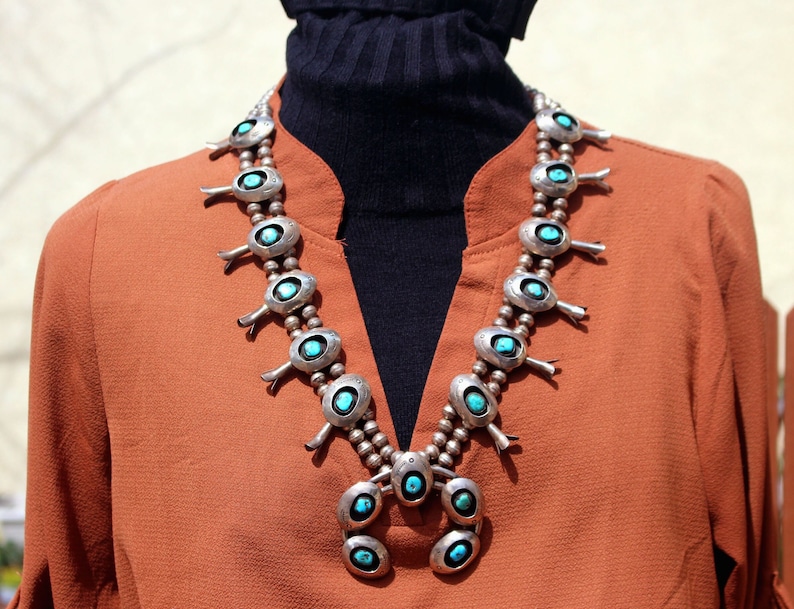 May include: A silver and turquoise necklace with a pendant design. The necklace is made of multiple oval-shaped turquoise stones with silver accents. The pendant is a cluster of smaller turquoise stones.