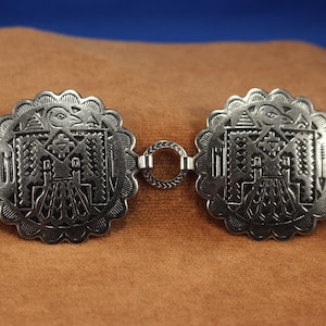 May include: A silver belt with two round, ornate, silver buckles. Each buckle features a detailed design of a stylized bird with geometric patterns.