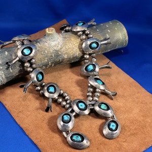 May include: A silver and turquoise necklace with a cluster of turquoise stones and silver beads. The necklace is on a brown wooden log and a brown suede surface.