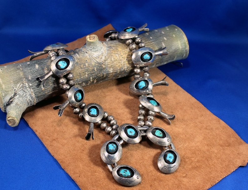 May include: A silver and turquoise necklace with a cluster of turquoise stones and silver beads. The necklace is on a brown wooden log and a brown suede surface.