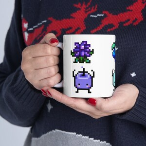 May include: White ceramic mug with a pixelated design featuring a purple flower, a purple creature with a single eye, and a blue creature with a single eye.
