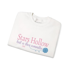 Könnte beinhalten: Gefaltetes weißes T-Shirt mit dem Text "Stars Hollow knit-a-thon committee 2006" in Rosa und Blau. Eine blaue Garnknäuel-Grafik ist ebenfalls abgebildet.