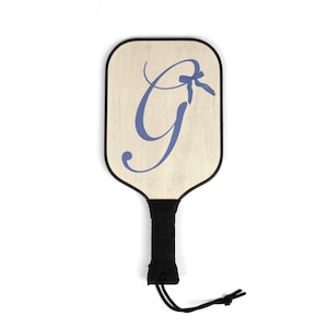 Op de afbeelding: Een houten paddle met een zwarte handgreep en een blauwe letter G met een blauwe strik erop. De paddle is voor het spelen van pickleball.