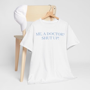 May include: White t-shirt with blue text that reads "ME, A DOCTOR? SHUT UP!"