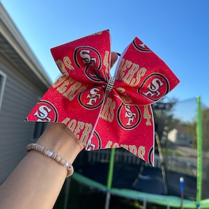 May include: A red and gold cheer bow with the San Francisco 49ers logo and the word "49ers" repeated on the bow. The bow has a white border and a rhinestone in the center.
