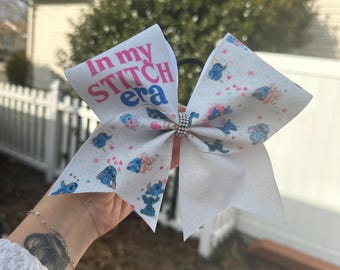 Puede incluir: Un lazo de animadora blanco con un estampado azul y rosa de personajes de Stitch de la película de Disney Lilo & Stitch. El lazo tiene un strass en el centro y el texto "In my Stitch era" impreso en la parte superior.