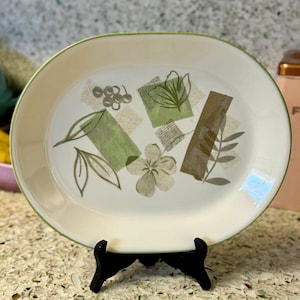 May include: A white oval serving platter with a green rim and a green and brown floral design. The platter is on a black stand.