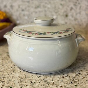 May include: Cream-colored ceramic casserole dish with a lid. The lid is decorated with a floral garland of pink roses and blue ribbons. The dish has two handles and a matching knob on the lid. The dish is sitting on a speckled surface.