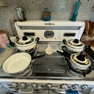 May include: A white vintage stovetop with four black and white floral patterned pots and a white ladle. The stovetop has four knobs and a black metal surface.
