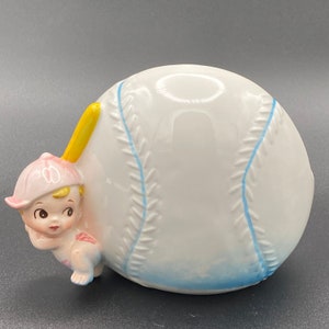 May include: A white ceramic baseball piggy bank with blue stitching and a small boy figurine holding a yellow bat. The boy is wearing a pink baseball cap and a pink and white outfit.