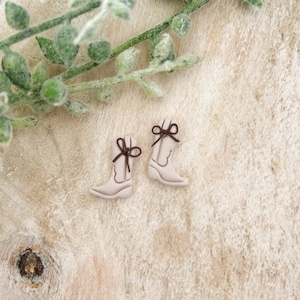 May include: A pair of small, light-colored cowboy boot earrings with dark brown bows. The earrings are set on a light wooden surface, with green leafy sprigs in the upper left corner.