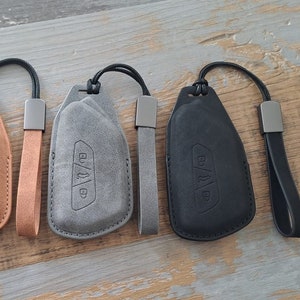 Puede incluir: Tres llaveros de coche en fundas de cuero marrón, gris y negro con correas de muñeca a juego. Cada funda tiene un símbolo de llave en relieve y un acento metálico. Los llaveros se muestran sobre una superficie de madera envejecida.