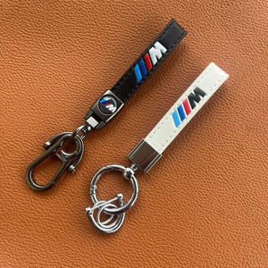 May include: Two keychains with the BMW M logo. One is black with a silver clasp, and the other is white with a silver keyring. Both have the M logo in blue, red, and white.