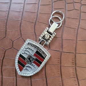 May include: Silver Porsche keychain with a shield-shaped emblem. The emblem features the Porsche logo, a black horse, and red and black stripes, all surrounded by sparkling crystals. The keychain is on a brown, textured surface.