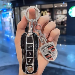 May include: A black car key fob and a matching Porsche keychain. The key fob has silver accents and a Porsche emblem. The keychain features the Porsche logo with red, black, and silver details, all encrusted with rhinestones.