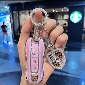 May include: A pink car key fob with a silver Porsche emblem and a matching keychain. The keychain has a shield-shaped charm with the Porsche logo and is encrusted with rhinestones. The key fob has buttons for locking and unlocking the car.