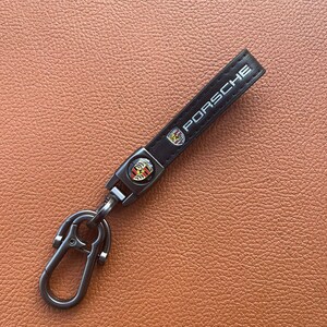 May include: Black leather keychain with the word "PORSCHE" in white lettering. The keychain has a metal clasp and a Porsche emblem. The background is a textured, brown surface.