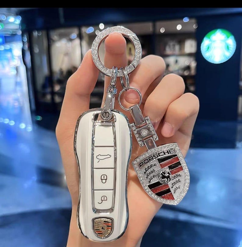 May include: White car key with a silver key ring and a Porsche emblem keychain. The key and keychain are adorned with sparkling rhinestones. The Porsche emblem features red and black accents.