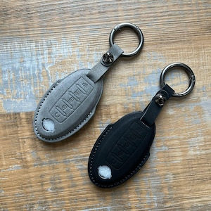 May include: Two leather key covers for car keys. One is grey and the other is black. Both have a key ring attached.