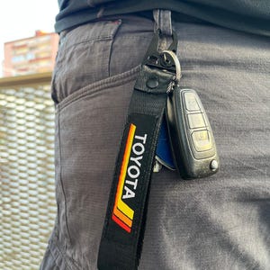 Peut inclure: Porte-clés noir avec un logo "TOYOTA" noir et jaune sur une sangle noire. Le porte-clés est attaché à un porte-clés argenté avec une clé de voiture noire.