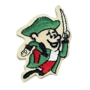 May include: Embroidered patch featuring a cartoon leprechaun in green and red attire, holding a sword. The leprechaun has a smiling face and is set against a cream-colored background. The patch is likely for clothing or accessories.