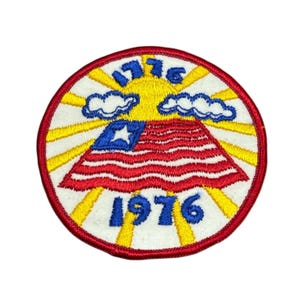 May include: Embroidered patch with a patriotic design. The patch features a red, white, and blue flag with a star, the year 1776 at the top, and 1976 at the bottom. The background includes a yellow sun and rays, with white clouds.