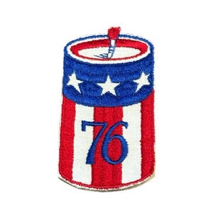 May include: Embroidered patch in the shape of a firecracker. The top is blue with white stars, the middle is red and white stripes, and the bottom has the number 76 in blue. A red wick is at the top.