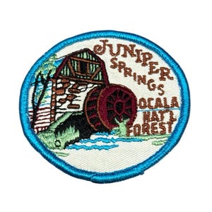 May include: Embroidered patch with a teal border featuring a water mill scene. The mill has a red water wheel and is set against a backdrop of green foliage and water. Text reads "Juniper Springs Ocala Natl Forest."