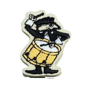 May include: Embroidered patch featuring a cartoon drummer in black and white attire, playing a yellow drum with black accents. The patch has a cream-colored background and is likely for clothing or accessories.