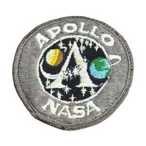 May include: A circular gray patch featuring the Apollo NASA logo. The logo includes a stylized letter "A" with a yellow and blue planet on either side, surrounded by stars. The words "APOLLO" and "NASA" are embroidered around the logo.