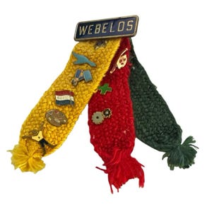 May include: A Webelos pin attached to three braided yarn strips in yellow, red, and dark green. The yellow strip features pins of a star, bird, flag, palette, and figure. The red strip has pins of a horn, cross, and gears. Each strip ends in a tassel.