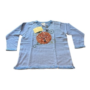 May include: A light blue long-sleeve sweater featuring a central embroidered orange pumpkin design with green vine accents and a small black spider. The garment includes a V-neckline and a tag attached near the collar area on the front side.