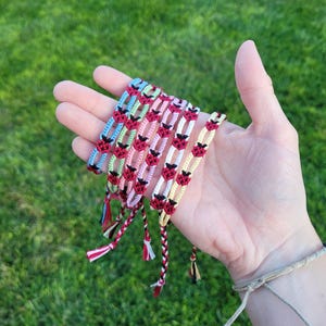 May include: A collection of handmade friendship bracelets in various colors, including blue, pink, and yellow. Each bracelet features a repeating pattern of red and black ladybug designs. The bracelets are displayed on a hand against a green background.