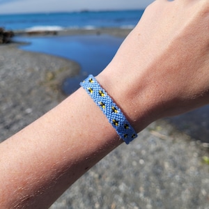 May include: A blue and yellow friendship bracelet with a geometric pattern. The bracelet is worn on a person's wrist.